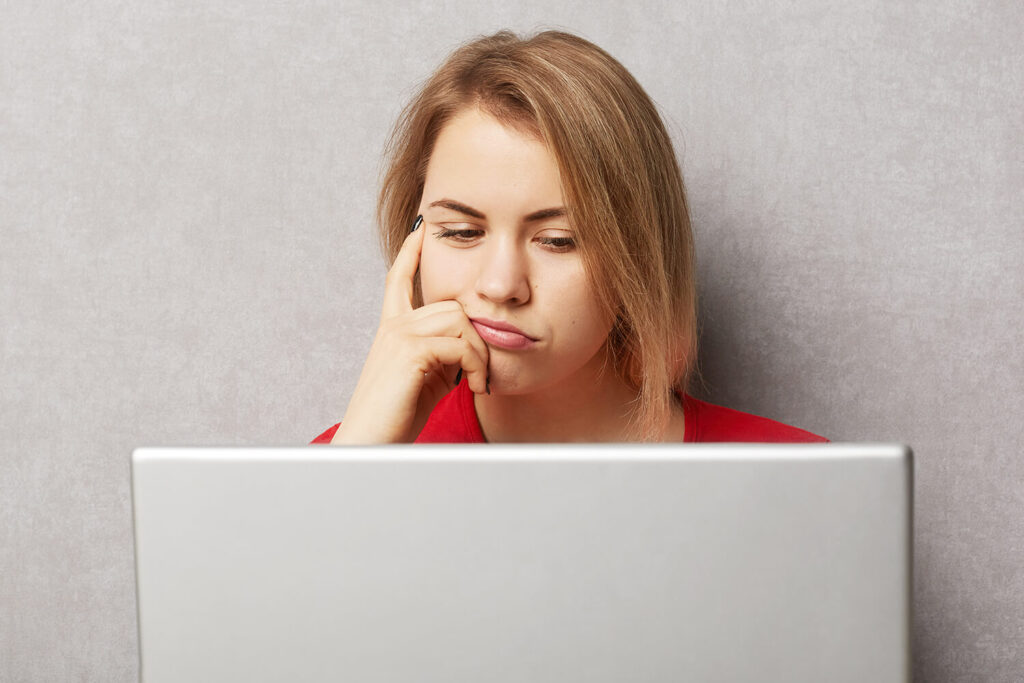 パソコンでオンライン・サイトの情報を読んでいる悲しげな顔をした若い白人女性の画像